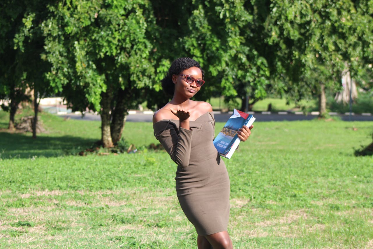 A confident woman poses with textbooks in a sunlit park, showcasing a vibrant academic spirit.